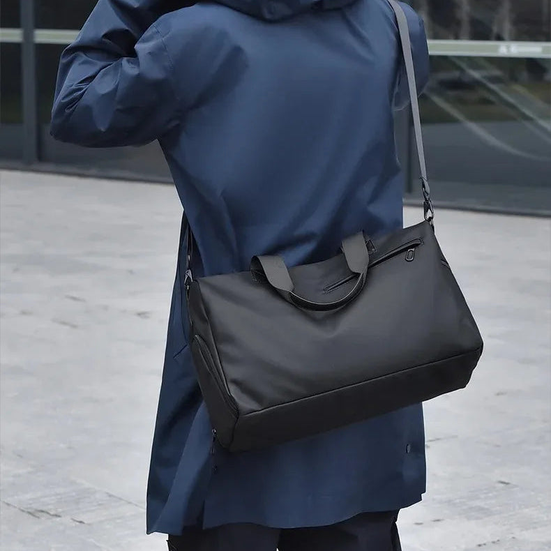 Black minimalist travel duffel bag with shoulder strap, worn by person in blue coat for urban commuting
