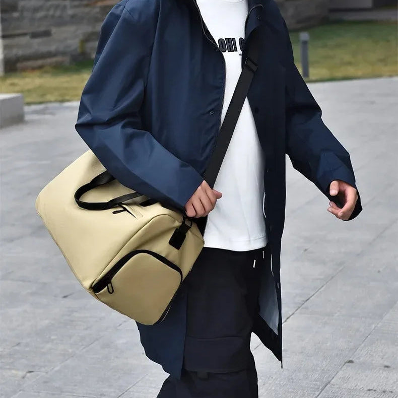 Beige minimalist travel duffel bag with black straps, worn crossbody by commuter in urban setting